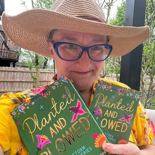 A person wearing a large straw hat, blue-framed glasses, and a yellow floral shirt holds two copies of the book 'Planted and Owed: Storm Stories' in front of their face. The photo is taken outdoors on a patio with trees and a wooden fence in the background.